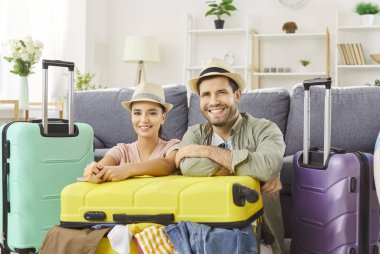 Gülümseyen kadın ve erkek çift seyahat için bavullarını topluyor, aile gezisine hazırlanıyor, kanepenin yanında, bavullarla çevrili bir şekilde oturuyorlar. Seyahat ve turizm konsepti.