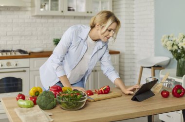 Modern mutfakta gülümseyen genç bir kadın tablete bakıyor, taze sebzeler hazırlarken online tarifi takip ediyor. Pozitif kadın video tarifi dersi izliyor ve sağlıklı yemek pişirme sürecinin tadını çıkarıyor..