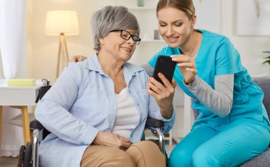 Tekerlekli sandalyedeki gülümseyen yaşlı kadın dost canlısı hemşireyle akıllı telefon kullanıyor. Neşeli kıdemli hasta, cep telefonunda bakıcıyla komik bir şey paylaşıyor. Bakımevi servisi ve hasta desteği.