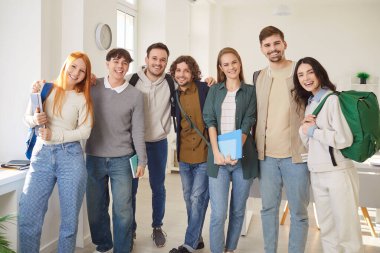 Modern sınıfta bir arada durup kameraya gülümseyen neşeli genç öğrencilerin grup portresi. Sırt çantalı ve defterli mutlu arkadaşlar birbirlerine sarılıyorlar, birlik ve dostluk gösteriyorlar..