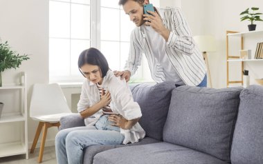 Endişeli genç koca, evdeki oturma odasındaki kanepede oturan kalp krizi geçiren genç kadın eşine cep telefonuyla acil ambulansı arıyor. Sağlık ve Tıp Konsepti.
