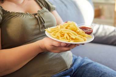 Aşırı kilolu bir kadının kanepeye uzanmış elinde patates kızartması ve ketçapla tabağı tutması. Şişman kadın fast food yiyor, lezzetli atıştırmalıkların tadını çıkarıyor. Sağlıksız yeme alışkanlıkları kavramı.