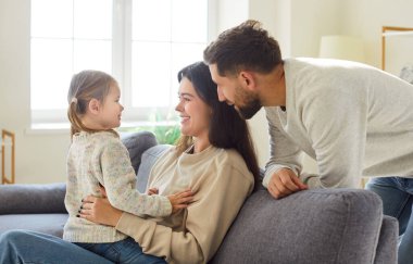 Üç kişilik mutlu bir ailenin portresi evdeki kanepede küçük tatlı bir kızla oturuyor ve gülümsüyor. Neşeli ebeveynler kızlarıyla vakit geçirmekten zevk alıyorlar. Aşk, ilgi ve mutlu aile kavramı.
