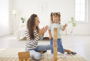 Mutlu genç anne, bebek bakıcısı küçük kıza tahta bloklardan oyuncak kuleler inşa etmede yardım ediyor, evde ısıtma katında çocukla oynuyor, dadılık yapıyor, annelik ve kreşe bakıyor.