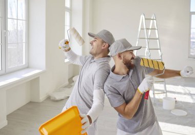 Üniformalı iki neşeli erkek ressam ellerinde mikrofon gibi boya ruloları tutuyorlar ve ferah bir odada şarkı söylüyorlar. Mutlu işçiler yenileme sırasında eğleniyorlar, iç dekorasyon sürecinin tadını çıkarıyorlar..