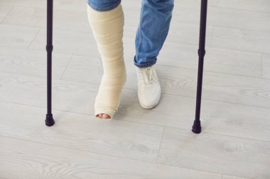 Oturma odasında koltuk değneklerini kullanarak yürüyen kırık bacaklı bir adamın kesilmiş fotoğrafı. Kazadan sonra askı ya da bandaj takan engelli bir erkek. İyileşme ve rehabilitasyon konsepti.