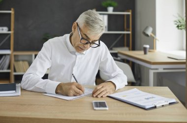 Ciddi bir kıdemli işadamı evrak işleriyle uğraşıyor ve kağıtlara not alıyor. Yetişkin bir iş adamının ofis masasında oturup bazı önemli rakamları yazması.