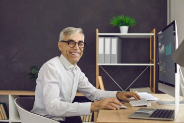 Ofiste gülümseyen profesyonel bir erkek, ekrandaki grafikleri analiz ediyor. Mutlu bir ofis çalışanının portresi, beyaz gömlek ve gözlük takıyor, modern bir iş yeri..