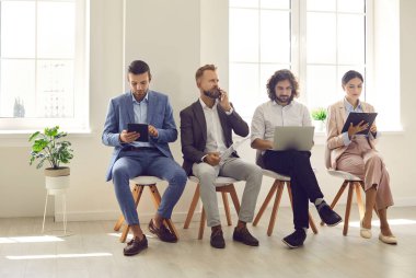 Çok ırklı genç çalışanlar şirket ofisindeki koltuklarda oturup şirketle görüşmeye hazırlanıyorlar. Motive olmuş çeşitli başvuru adayları cihaz araçları kullanarak işe alım konuşmasına hazırlanıyor.