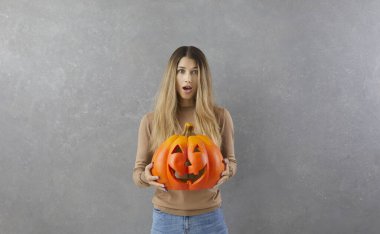 Elinde balkabağıyla duran komik, şaşırmış ve korkmuş genç bir kızın portresi. Gri arka planda duran Cadılar Bayramı balkabağıyla poz veren sıradan elbiseli bir kız. Cadılar Bayramı konsepti Pankart.