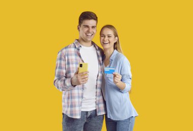 Aşırı neşeli beyaz bir çift telefon ve kredi kartı tutuyor ve internet üzerinden internet siparişi için gülümsüyor sarı stüdyoda birbirlerine sarılıyorlar.