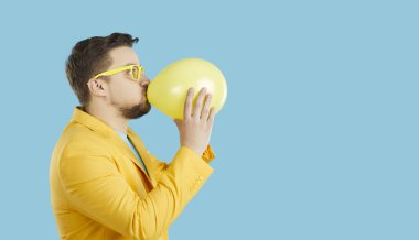 Mavi stüdyo arka planında hava balonu şişiren genç beyaz adamın yan görüntüsü. Hava balonlu adamı geniş açıyla çek. Eğlence ve eğlence. Boşluğu kopyala, boş reklam yerleştirmesi.