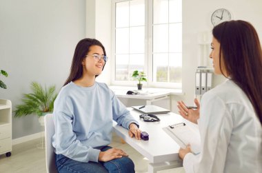 Doktor, muayenehanede parmak ucu nabız oksimetresi kullanan genç bir hastayla konuşuyor. Gülümseyen kadın klinikteki muayene sırasında masada oturan doktoru dinliyor. Tıp konsepti.