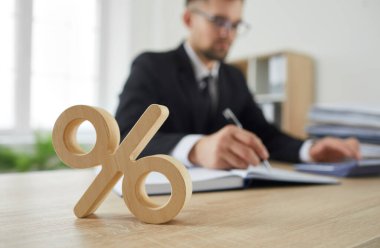 Şirket vergisi ve faiz oranı sembolü olarak masadaki ahşap yüzde işaretini kapat. Takım elbiseli, hesap makinesinde hesap yapan ciddi meşgul bir adamın arka planında duran yüzde işareti. Bulanık arkaplan.