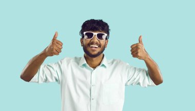 Genç etnik adam yaz tatilinin tadını çıkarıyor ve harika vakit geçiriyor. Tişörtlü ve modern güneş gözlüklü yakışıklı Hintli adamın portresi. Kameraya bakıp mutlu yüz ifadesi takınıyor ve baş parmağını kaldırıyor.