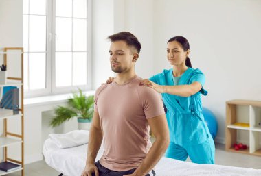 Modern fizik tedavi merkezinde kadın fizyoterapist erkek hastalara rehabilitasyon egzersizleri yaptırıyor. Doktor omuzlarına masaj yaparken rahat genç bir adam muayene odasında oturuyor.