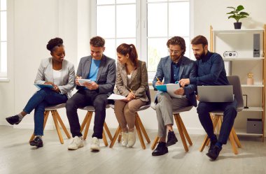 İş yerindeki adaylar iletişim kurup görüşmeye hazırlanıyorlar. Elinde belgelerle gülümseyen kadın ve erkekler sırayla koltuklarda oturuyorlar. İstihdam kavramı.