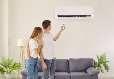 Oturma odasında klimayı açan kiracılar temiz hava soluyor. Erkek ve kadın kiracılar evde durum cihazı kullanıyorlar. İç mekan havalandırması ve modern teknoloji.
