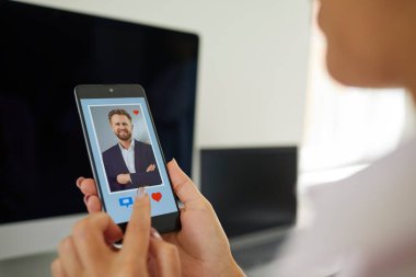 İnternette aşkı bulmak. Çevrimiçi çöpçatanlık sitesinde ya da mobil uygulama sitesinde genç ve çekici iş adamının fotoğrafı altında kırmızı kalp ve mesaj düğmeleri olan telefon ekranına bakan yakın plan kadın