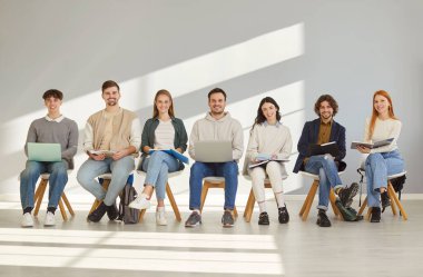 Genç erkekler ve kadınlar gündelik giysiler içinde sıra sıra sıra oturup röportaj davetiyesi bekliyorlar. İş adayları gri duvar arkasında oturuyorlar. İşe alım konsepti.
