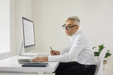 Modern ofiste çalışan olgun muhasebeci iş adamı. Gözlüklü ciddi bir beyaz adam dizüstü bilgisayar ve bilgisayarla masada oturuyor, elektronik iş kağıtlarıyla çalışıyor ve notlar alıyor.