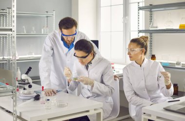 Laboratuvarda yeni bir bilim adamı keşfi. İki kadın ve bir erkek deney tüpleri ve mataralarla araştırma yapıyorlar. Beyaz önlüklü ve gözlüklü çalışanlar. Ciddi bir adam ve kadın kimyasal örneğe bakıyor..