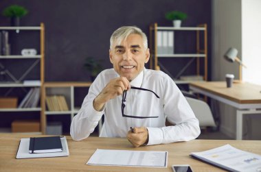 Ofis masasında oturan, elinde gözlük olan ve kameraya bakan son sınıf öğrencisinin portresi. Olgun iş adamı, CEO, yönetici, finans danışmanı, iş adamı ya da İnsan Kaynakları müdürünün webcam görüntüsü