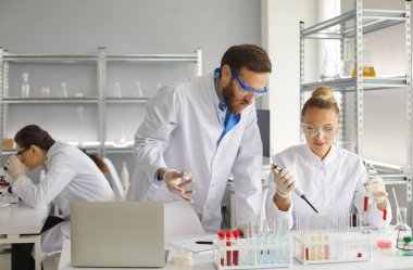 Kadın ve erkek bilim adamları laboratuarda çalışırken örnekleri analiz etmek için birlikte çalışırlar. Deneyimli bilim adamlarından oluşan bir ekip birkaç test tüpünden birine sıvı eklemek için bir pipet kullanır. Tıp konsepti.