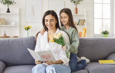 Gülümseyen bir kız çocuğunun portresi mutlu genç anneyi tebrik ediyor bir buket lale ve tebrik kartıyla evdeki kanepede oturuyor. Uluslararası Kadınlar ya da Anneler Günü konsepti.