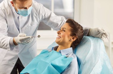 Modern diş hekimliği kliniğinde gülümseyen güzel bir kadının dişlerini diş aletleriyle muayene edip tedavi eden genç bir dişçi. Diş sağlığı, hijyen ve tıp konsepti.