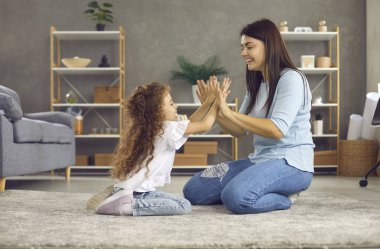 Neşeli genç anne, küçük tatlı kızının ellerini çırpmasıyla eğlenceli oyunlar oynuyor. Sevgi dolu aile evde oturma odasında yerde otururken eğleniyor. Çocukluk ve annelik kavramı.