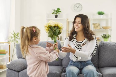Çocuk kızı, evdeki oturma odasındaki kanepede oturan mutlu genç annesini tebrik ediyor. Küçük tatlı kız, anne ve babasına çiçek veriyor. Anneler Günü konsepti.