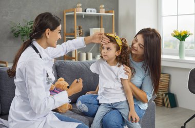 Kadın çocuk doktoru, ev ziyareti sırasında küçük bir kızın ateşini ölçüyor. Dost canlısı doktor elini annesinin kollarında oturan bir çocuğun alnına koydu..