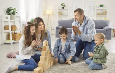 Üç çocuğuyla eğitim oyunu oynayan mutlu ebeveynler, oturma odasındaki yumuşak halıda birlikte tahta küplerle oyuncak kule inşa ediyorlar. Evde ve çocuk yaratıcılığında aile aktiviteleri.