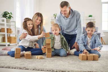 Evde mutlu büyük bir aile, anne, baba ve neşeli sevimli çocuklar halının üzerinde oynuyorlar, tahta küpleri istifliyorlar, oyuncak yapı taşları inşa ediyorlar, gülümsüyorlar, eğleniyorlar, eğleniyorlar, eğleniyorlar, eğleniyorlar.
