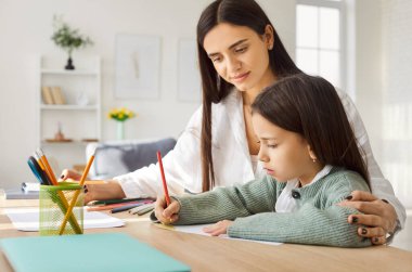 Bakıcı anne, parlak odadaki renkli kalemlerle yazarken ya da çizerken küçük kızına rehberlik ediyor. Çocuk görevine odaklanırken anne ona sarılıyor ve öğrenme sırasında destek oluyor..