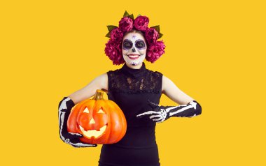 Geleneksel Ölüler Günü kostümü giymiş bir kadın işaret parmağıyla balkabağını işaret ediyor. Kafasında şeker kafatası ve çiçek çelengi olan neşeli kadın turuncu arka planda parlayan balkabağı tutuyor..