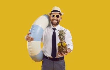 Mutlu ofis çalışanı yaz tatilinin tadını çıkarıyor. Beyaz gömlekli, siyah kravatlı, güneş gözlüklü, güneş şapkalı, elinde su halkası olan gülümseyen genç bir adam ve sarı arka planda izole edilmiş komik bir ananas.