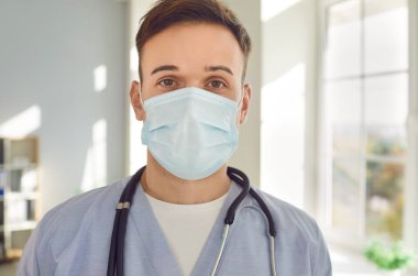 Tıbbi maske takan genç bir doktor portresi ve steteskoplu mavi üniformalı hastalara klinikte yardım etmek için kameraya bakıyor. İlaç ve sağlık hizmetleri kavramı.