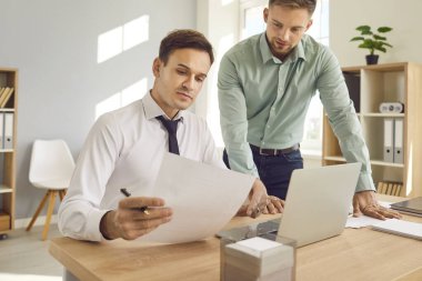 Ofiste çalışan iş adamları. İki ciddi adam birlikte kağıt okuyorlar. İki erkek arkadaş evrak işi yapıyor ve dizüstü bilgisayar kullanıyor.