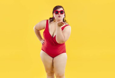 Stüdyo portresi, kırmızı mayo ve güneş gözlüğü takan şişman, güzel bir kadının sarı arka planda izole edilmiş kalçasında duran ve hava öpücüğü veren bir kadın.