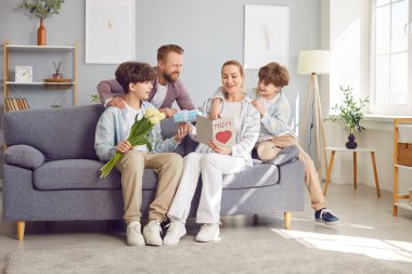 Mutlu baba ve Clildren, iki oğul, Uluslararası Kadınlar Günü, doğum günü ya da Anneler Günü ile annemi kutluyorlar. Gülümseyen anne, sevgi dolu ailesinden kart, hediye kutusu ve bahar çiçekleri alıyor.