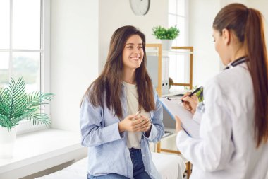 Beyaz önlüklü pratisyen hekim, klinikteki genç bayan hastaya profesyonel danışmanlık hizmeti veriyor, hastane ziyareti, hastalık tedavi planı hakkında bilgi veriyor, panoya yazılan ilaçları anlatıyor.