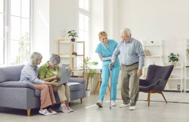 Huzurevinin oturma odasında bir grup mutlu yaşlı insan. Hemşire yaşlı bir adamın bastonla yürümesine yardım eder. Yaşlılar için konsept bakım, huzurevi, emekliler için boş zaman, yaşlı ebeveynler