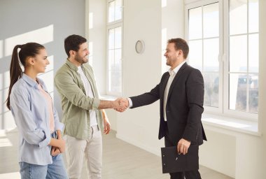 Mutlu gülümseyen genç müşteriler profesyonel bir emlakçıyla el sıkışıyor ve yeni bir dairede emlak almak için anlaşma yapıyorlar. Aile ev sahibi olmaya hazır. Emlak piyasası kavramı