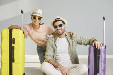 Plaj şapkalı ve güneş gözlüklü mutlu bir çift gri stüdyo arka planında bavullarıyla oturuyor. Gülümseyen adam ve kadın tatil için hazır. Tatil ve yaz yolculuğu konsepti.