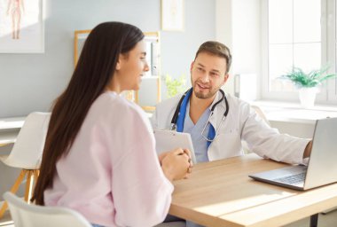 Beyaz üniformalı gülümseyen erkek doktor laptopta sağlık sonuçlarını gösteriyor, kadın hastaya danışıyor, el sıkışıyor, erkek pratisyen hekim modern hastanedeki kadın müşteriyle konuşmaktan mutlu oluyor.