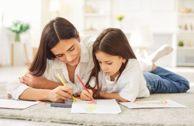 Neşeli gülümseyen annenin beyaz bir kağıda renkli kalemlerle çizdiği ve genç kız çocuğunun oturma odasında yerde yattığı yakın plan. Aile eğlencesi ve eğitim kavramı.