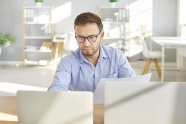 Resmi tişört ve gözlük takan kendine güvenen genç iş adamının portresi çalışma masasında oturuyor, modern ofiste bir dizüstü bilgisayarda çalışıyor ve bilgisayar monitörüne bakıyor..