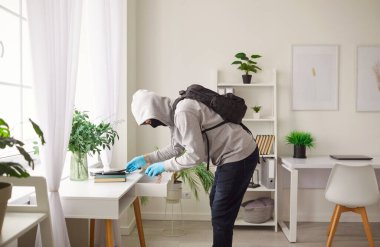 Maskeli erkek hırsız ya da soyguncu evde hırsızlık yapıyor, masadan değerli şeyler çalıyor. Güvenlik riskleri, tehlike ve suç faaliyetlerini gösteren suç mahalli, ev güvenliği kaygılarını vurguluyor.